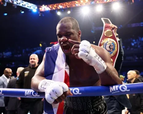 Daniel Dubois celebrates victory after the WBO world heavyweight title fight against Fabio Wardley in the ring with a championship belt visible behind him, draped in a union jack